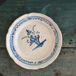 Plat rond en faïence dit cul noir, réalisé vers 1850, au décor bleu cobalt peint à la main motif un panier fleuri stylisé et bordure guirlande inspiration lambrequins. Cette pièce typique des faïences rurales françaises du XIXᵉ siècle évoque les productions de Sinceny ou Ger, connues pour leur émail craquelé et leurs décors bleus d’une grande fraîcheur