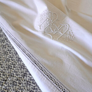 Drap ancien en lin élément d’un trousseau de mariage vers 1900–1920. Grande taille, avec monogramme brodé à la main "L A" entouré de motifs floraux plumetis. La bordure est ornée d’une broderie ajourée point Venise à fils tirés réalisée à la main, posé sur une courte pointe fleurie matelassée en coton